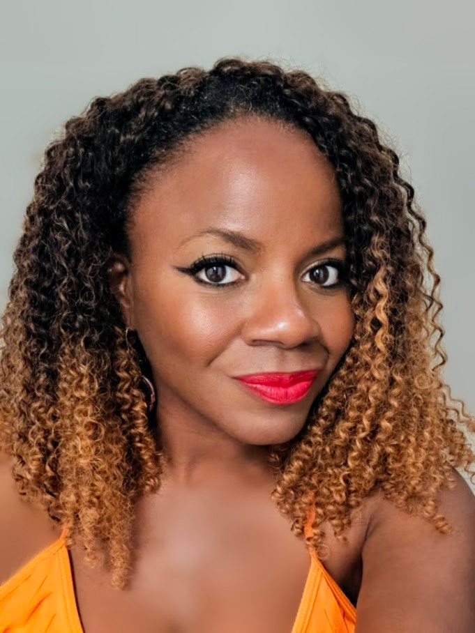 A portrait of a person with curly, two-toned hair wearing an orange top and bright red lipstick against a gray background.