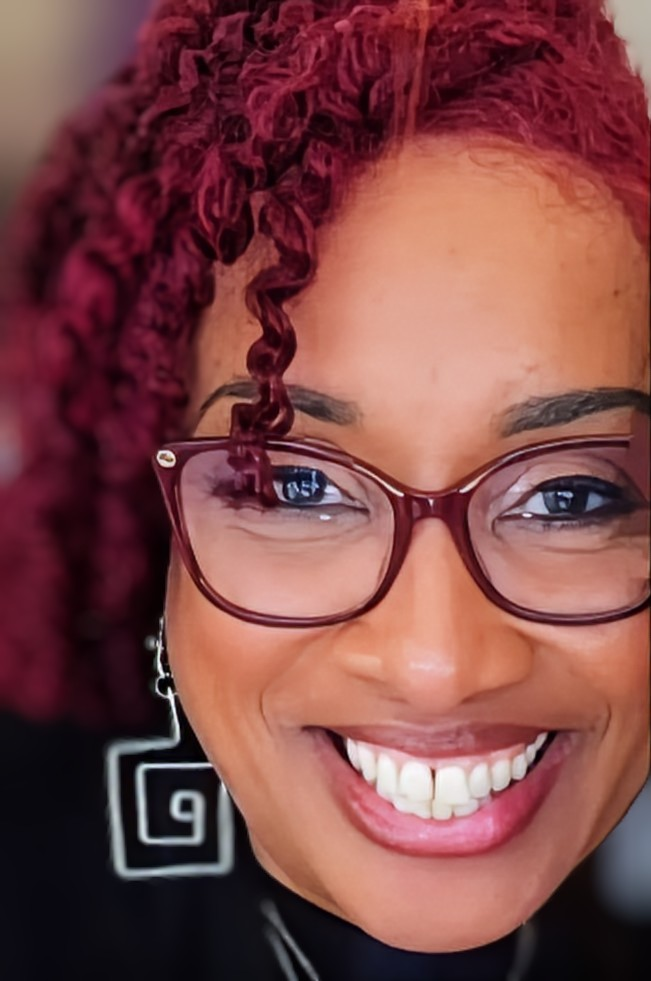 A smiling person with bright magenta curly hair and geometric earrings, wearing dark-framed glasses.