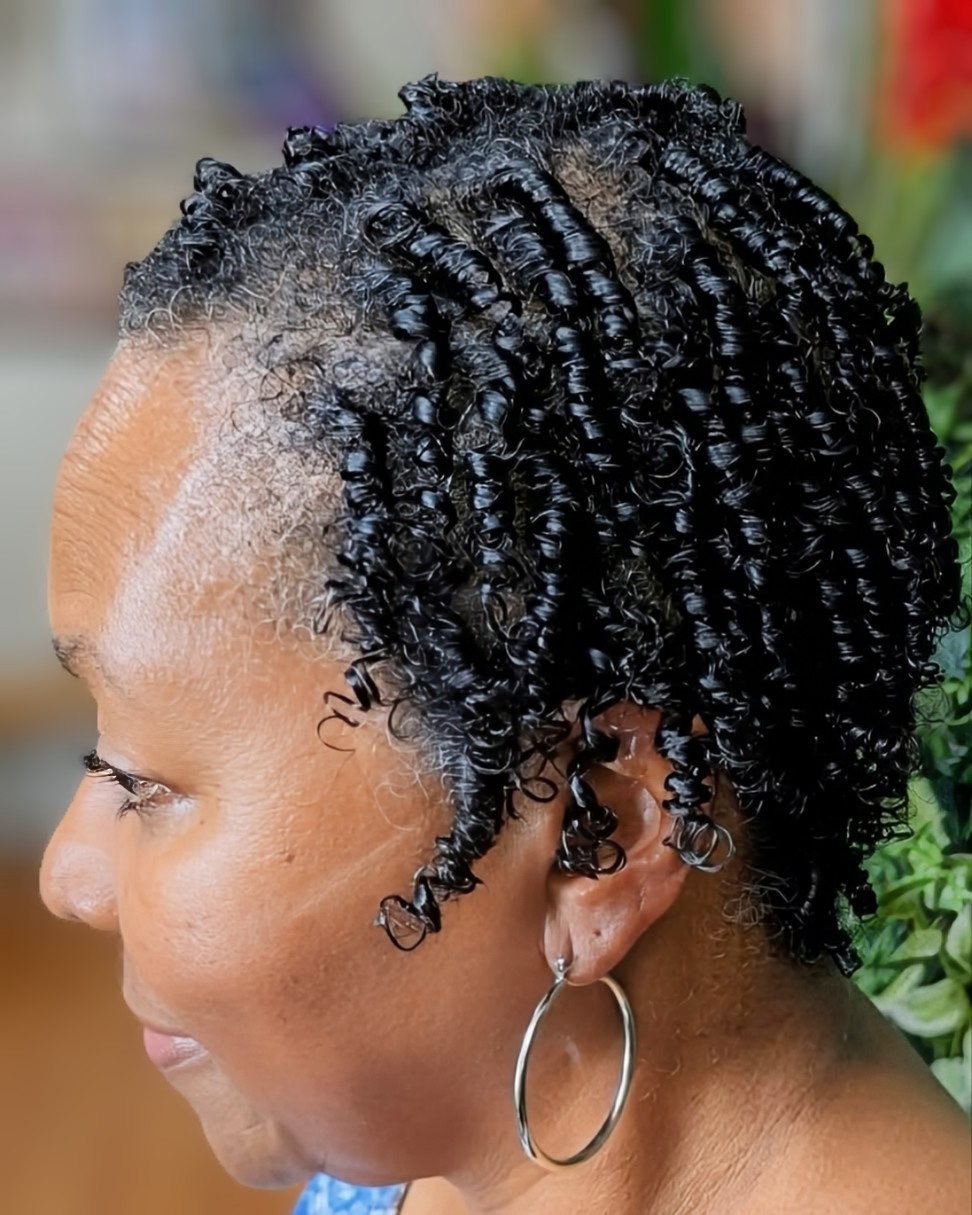 Profile view of a person with short, dark hair styled in defined, uniform finger coils, wearing large silver hoop earrings.