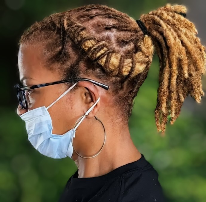 Side profile of a person with blonde dreadlocks styled in a ponytail, wearing glasses and a light blue face mask.
