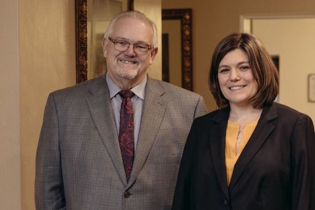 Two people smiling indoors, standing side by side in business attire.