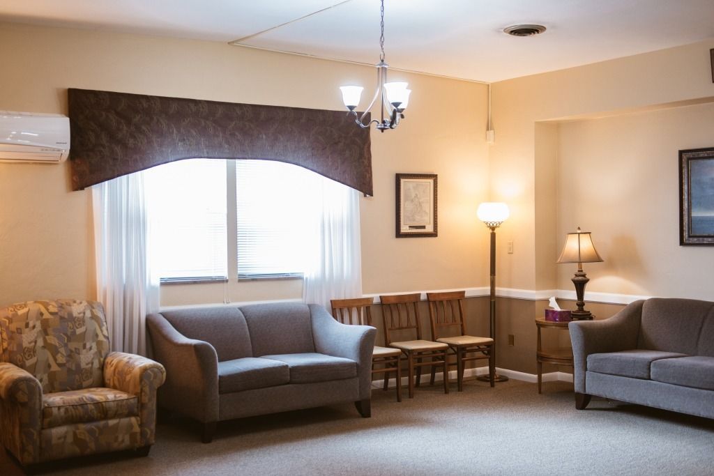 Living room with two gray sofas, an armchair, a small table, lamp, and window with dark valance