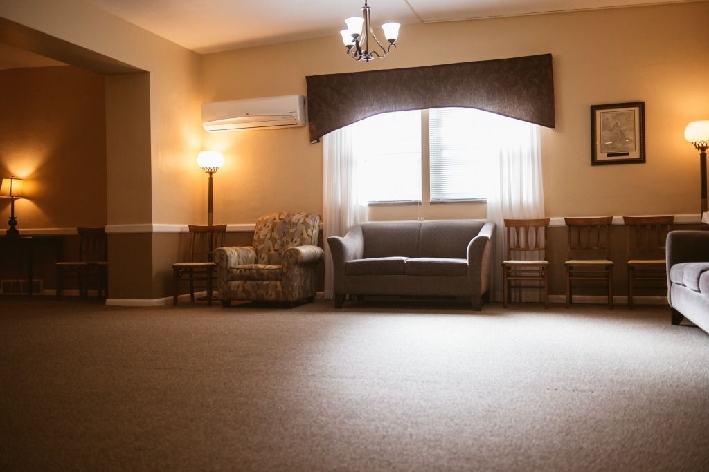 Empty waiting room with beige carpet, chairs along the walls, and a window with curtains in warm lighting