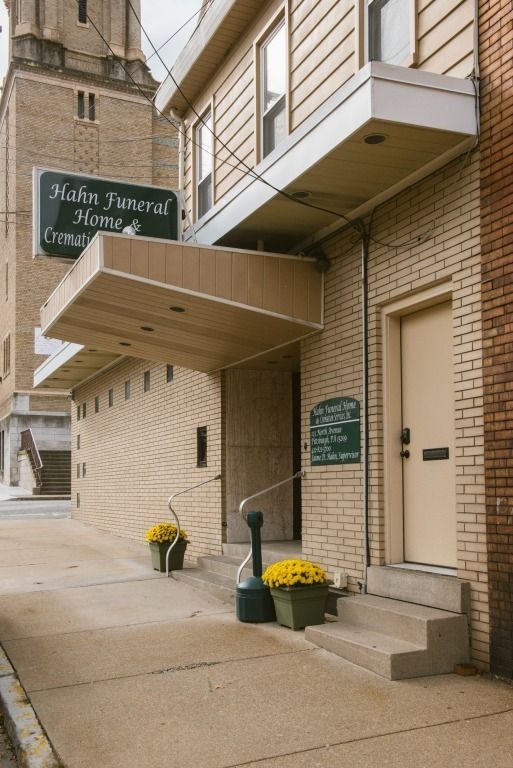 Brick building entrance with awning, green funeral home sign, and yellow potted flowers on the sidewalk