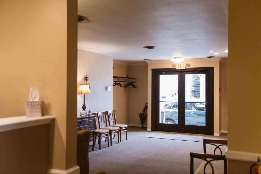 Empty beige lobby with chairs, lamp, carpet, and glass doors opening to a bright outdoor patio