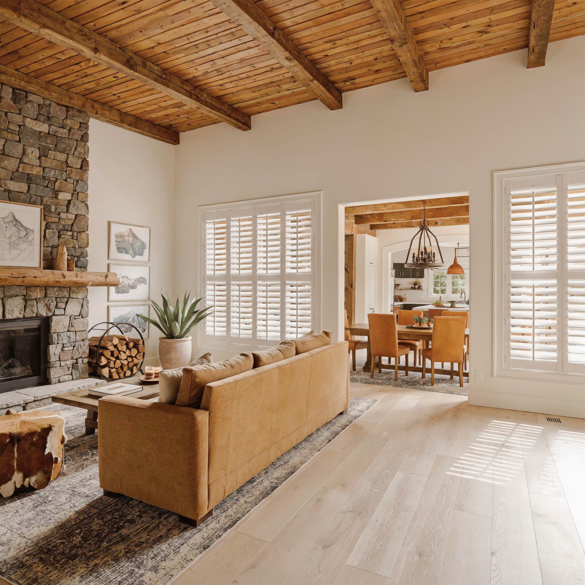Bright living room with stone fireplace, tan sofa, white shutters, and wooden ceiling beams