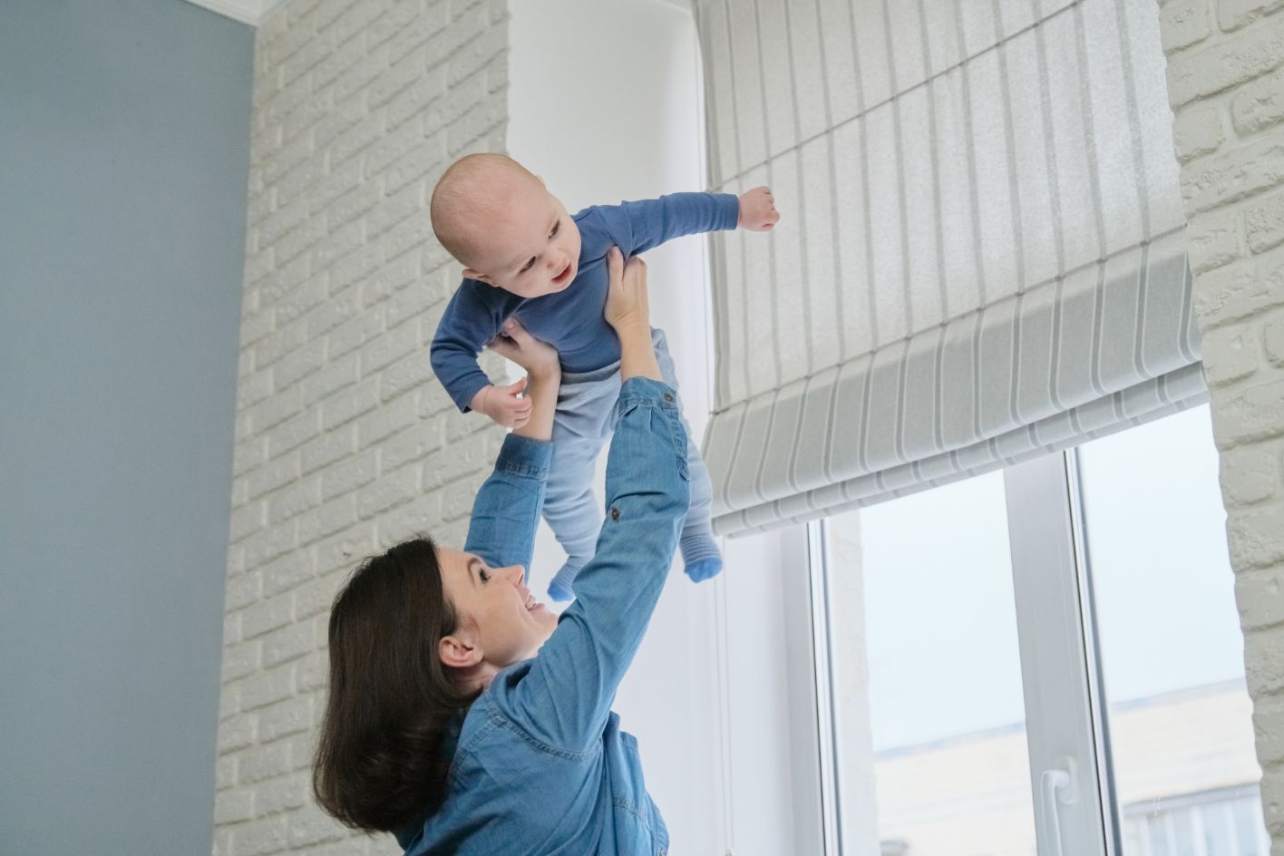 Woman holding a baby up in the air near a window. The baby is smiling, and the woman is looking up.