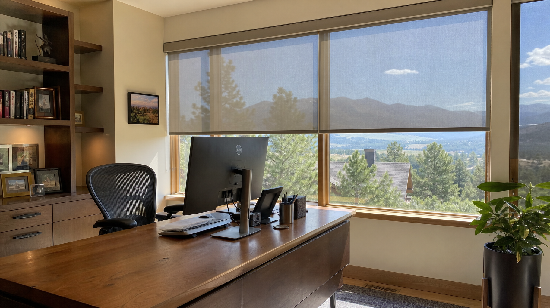 Modern office with wooden desk, computer, bookshelves, and large window with solar shades overlooking mountains and water