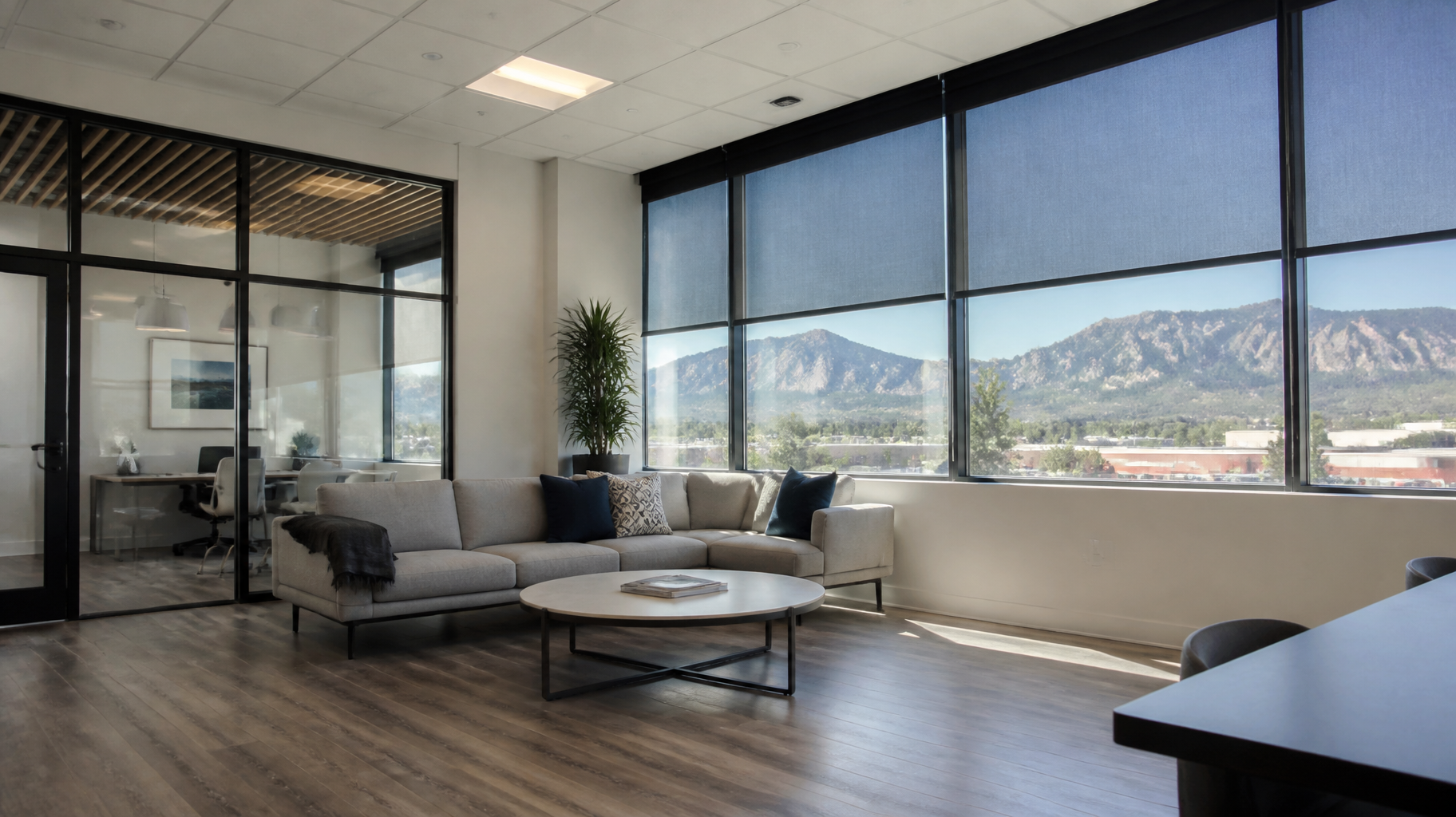 Modern office lounge with gray sofas, round coffee table, wood floors, and large windows with solar shades.