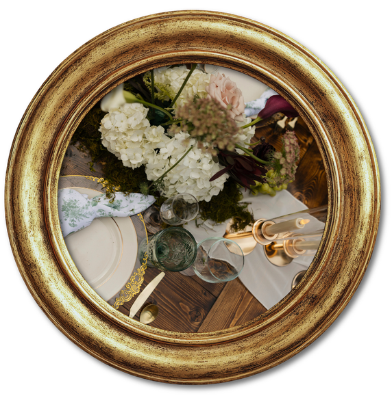 Round gold-framed mirror reflecting a white floral arrangement and a table setting with glassware and candles