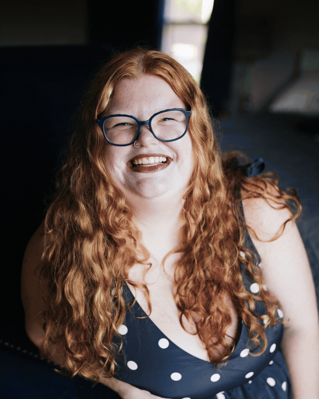 Smiling woman with red curly hair and glasses in a navy polka-dot dress, seated against a dark background