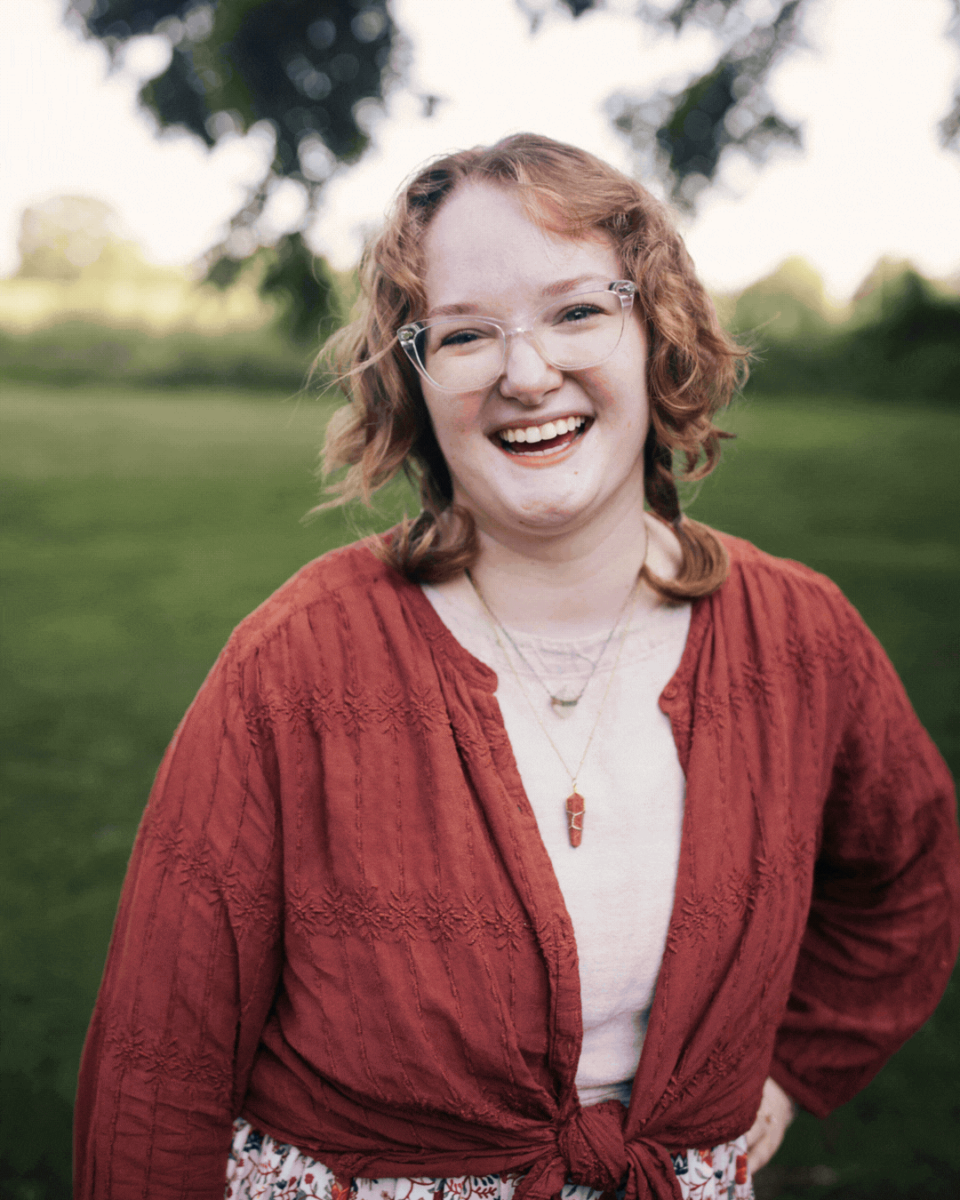 Smiling person in a red top standing outdoors on a grassy field