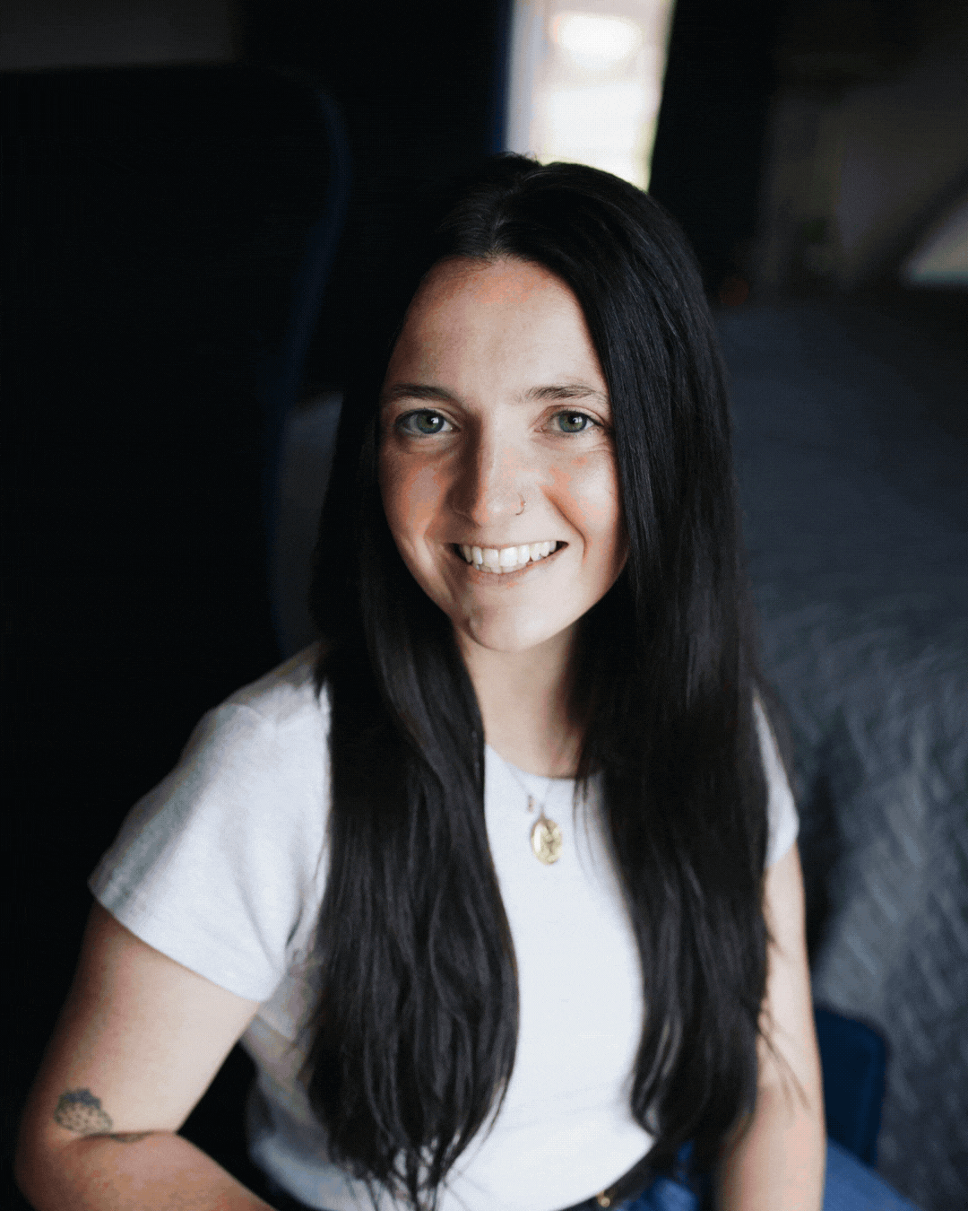 Smiling person with long black hair wearing a white shirt, seated on a dark couch indoors