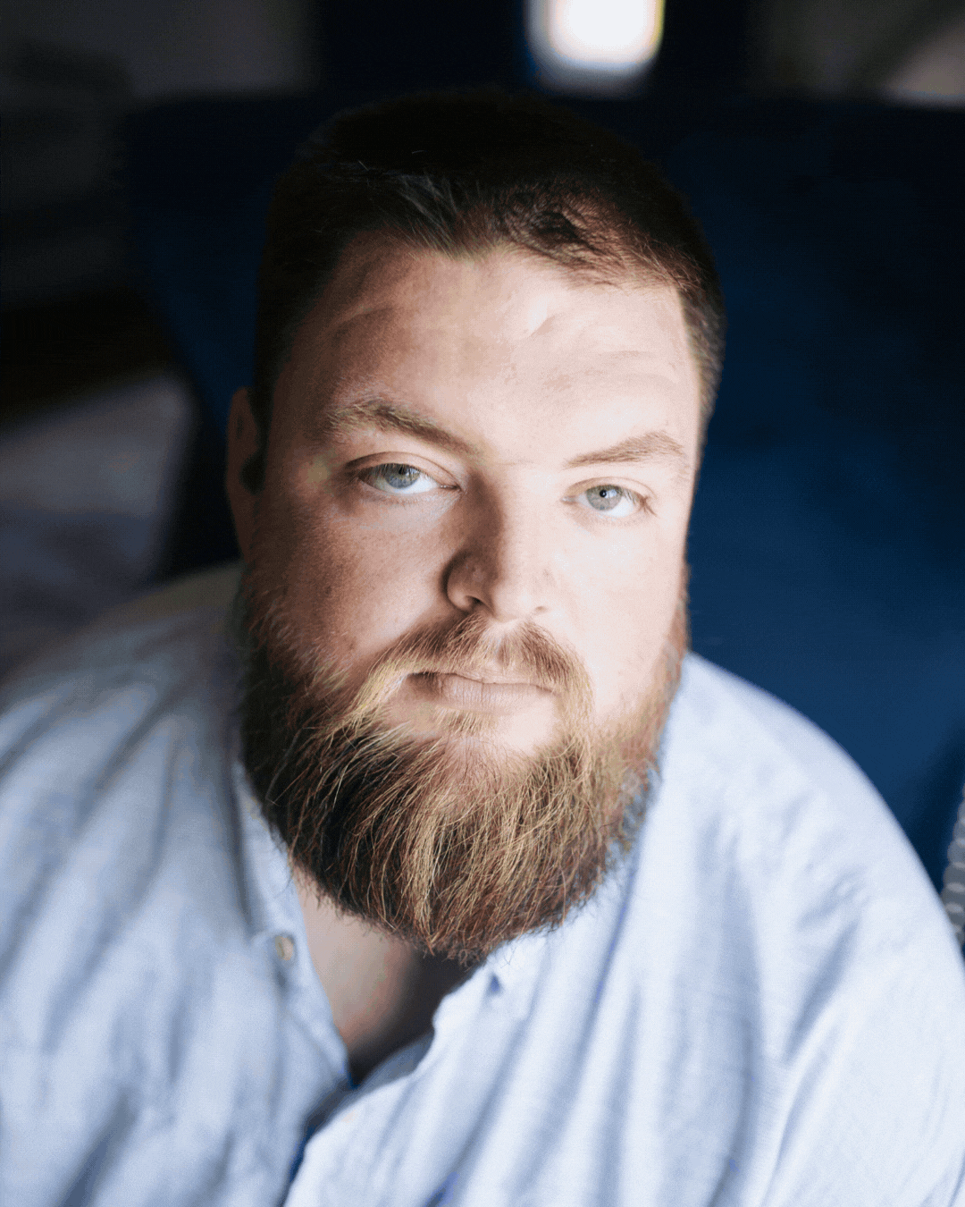 Close-up portrait of a bearded man in a light shirt, looking at the camera against a dark background