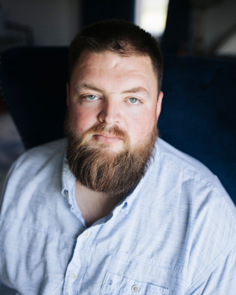 Close-up portrait of a bearded man in a light blue shirt, looking at the camera against a dark blurred background