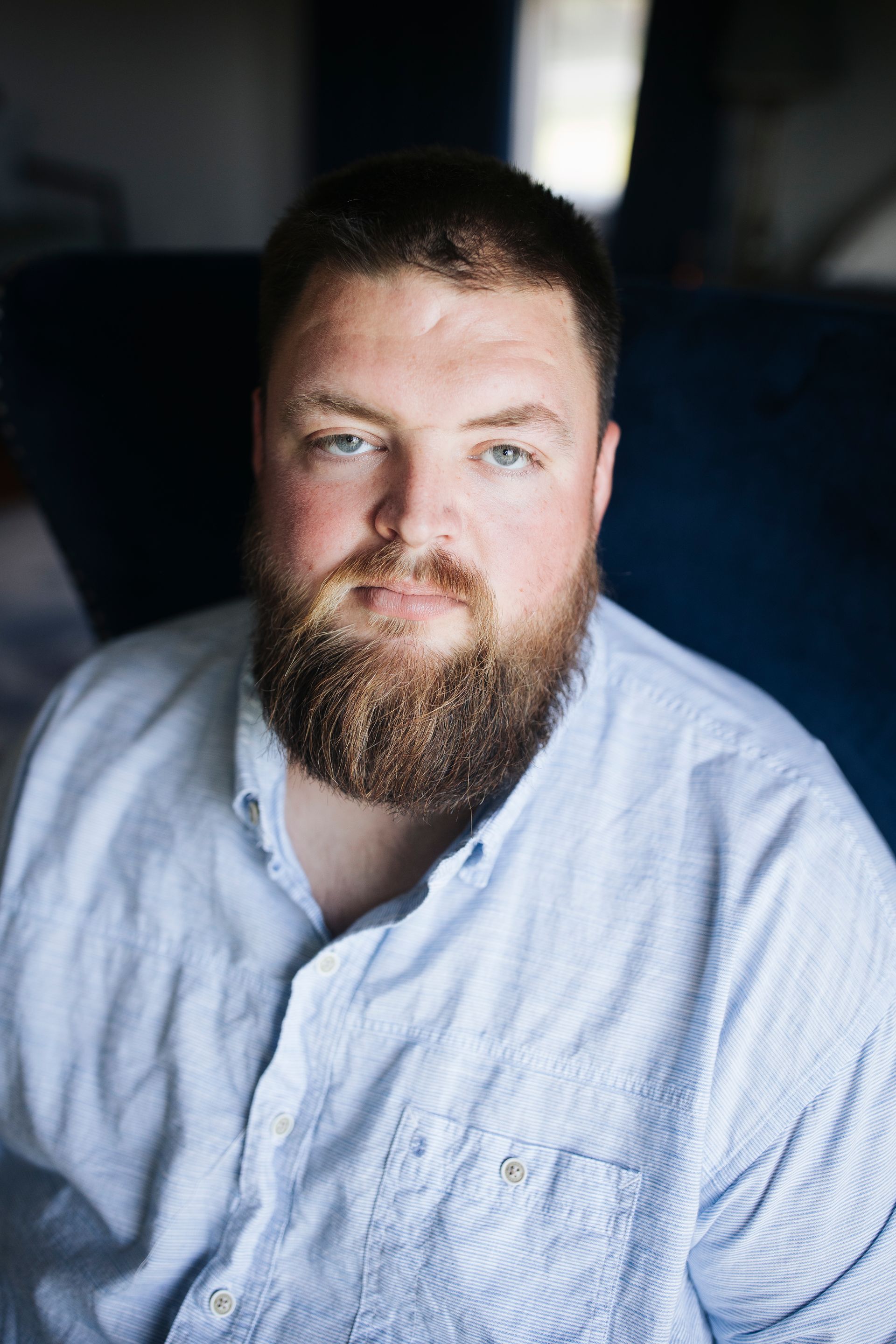 Close-up portrait of a bearded man in a light blue shirt, looking at the camera against a dark blurred background