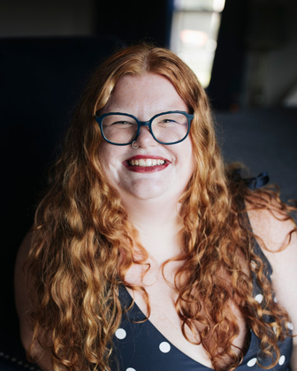 Smiling woman with red curly hair and glasses in a black polka-dot top