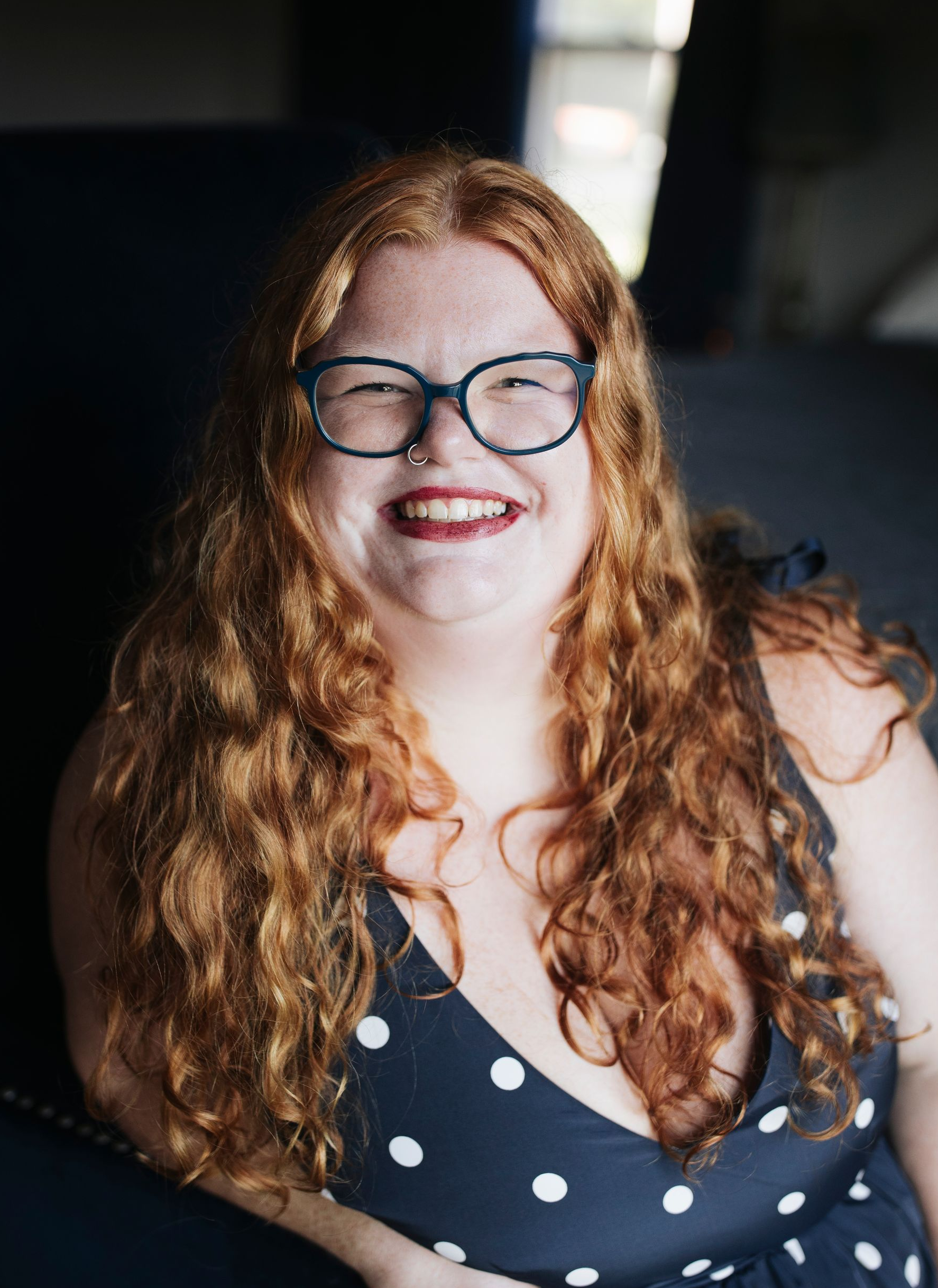 Smiling woman with red curly hair and glasses in a black polka-dot top