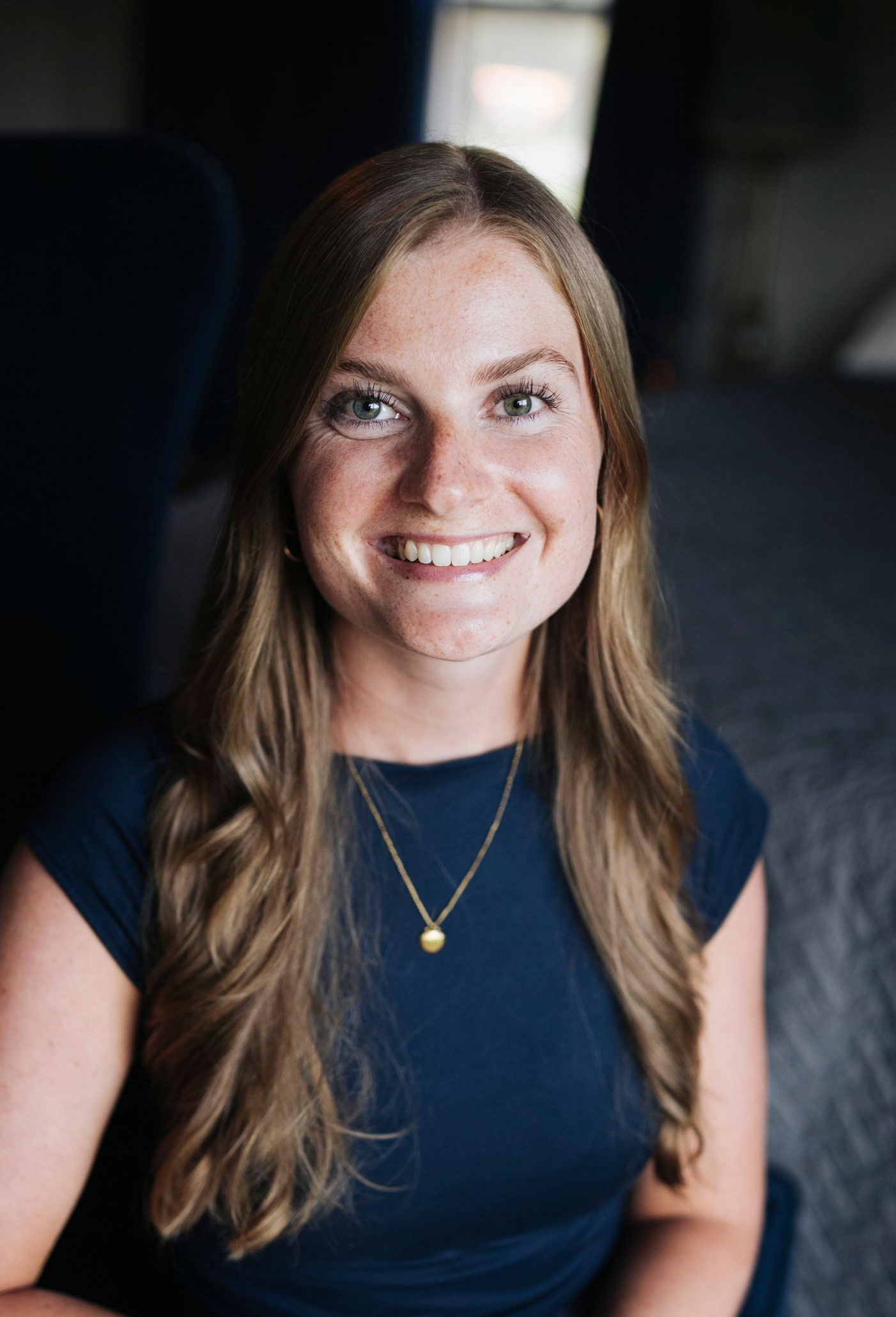Smiling person with long hair wearing a navy shirt and gold necklace against a blurred dark background