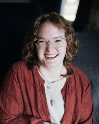 Smiling person with glasses and curly hair, wearing a red jacket, against a dark background