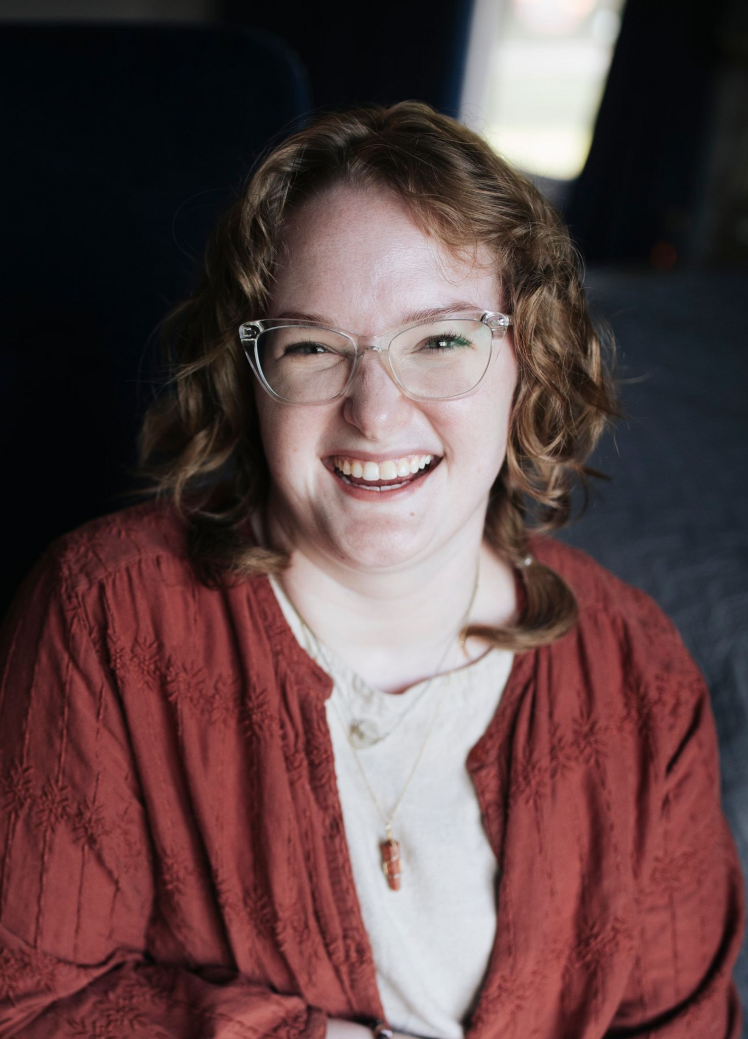 Smiling person with glasses and curly hair, wearing a red jacket, against a dark background