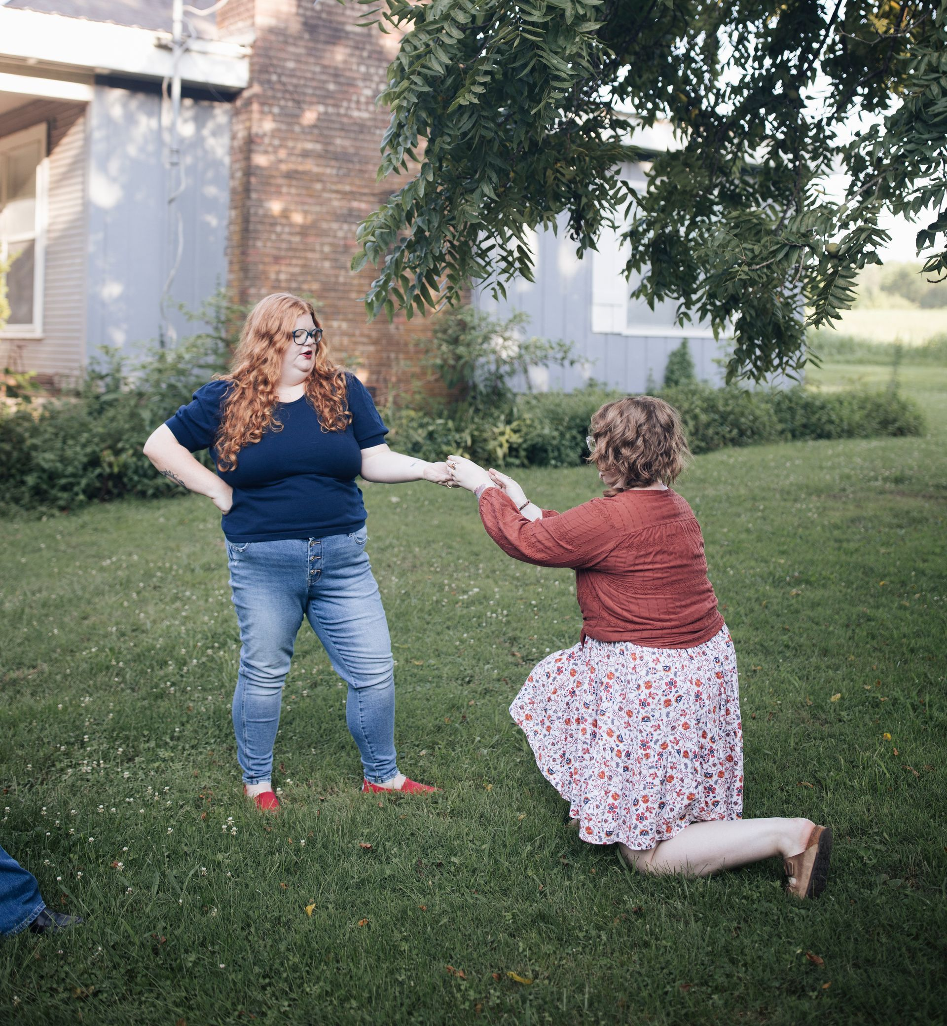 Woman kneeling on grass, holding hands with a standing person near a brick house in a yard