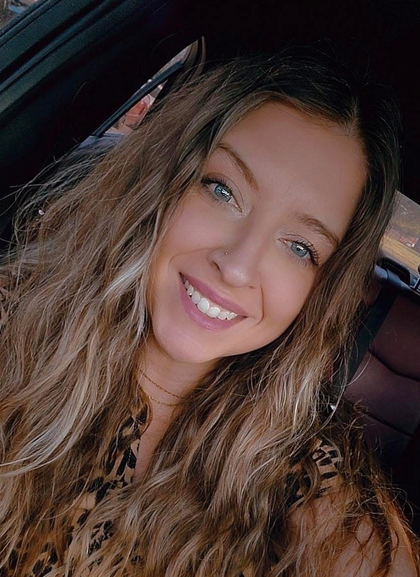 Smiling person with long wavy hair sitting in a car, wearing a patterned top