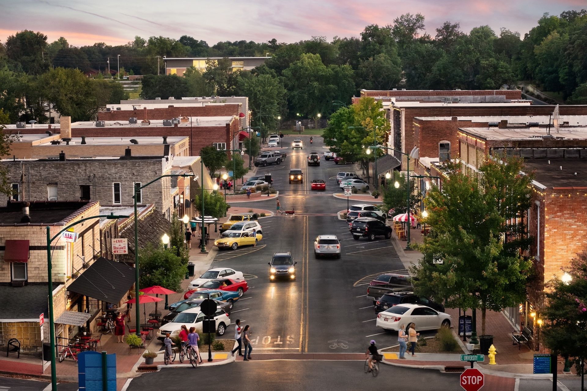 Downtown street at dusk with cars, pedestrians, and brick buildings under a pink sky