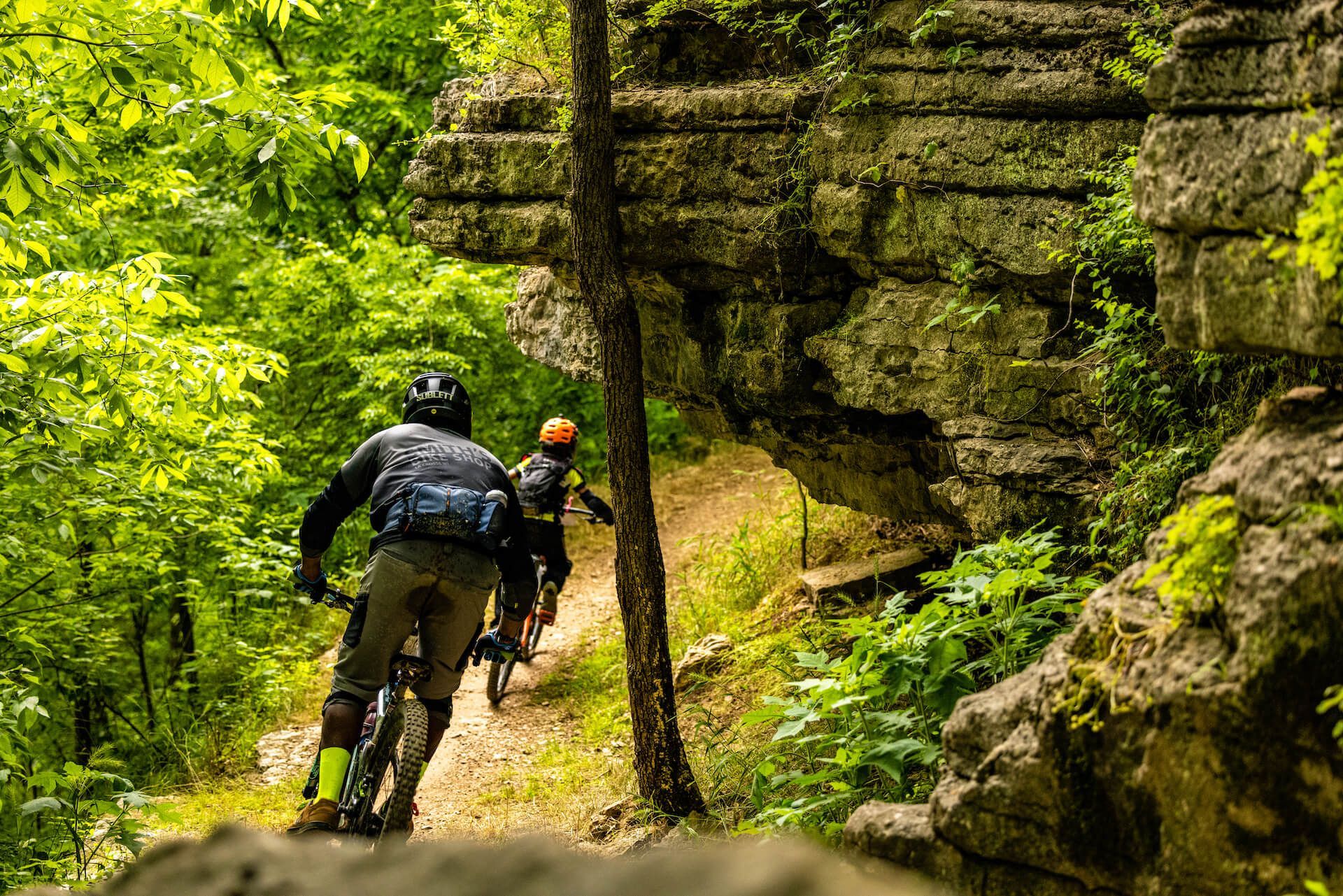 Two mountain bikers ride along a narrow forest trail beside mossy rock cliffs.