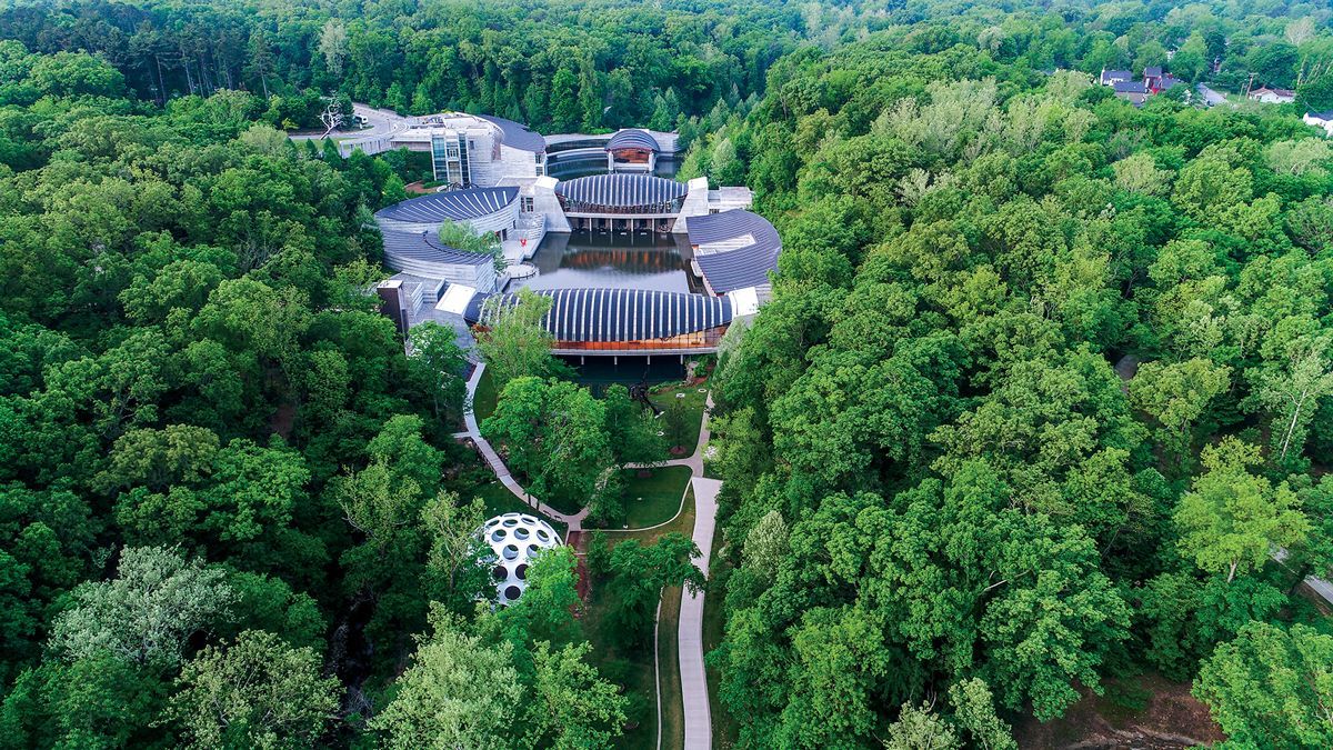 Aerial view of a large building complex surrounded by dense green forest, with a road leading in.