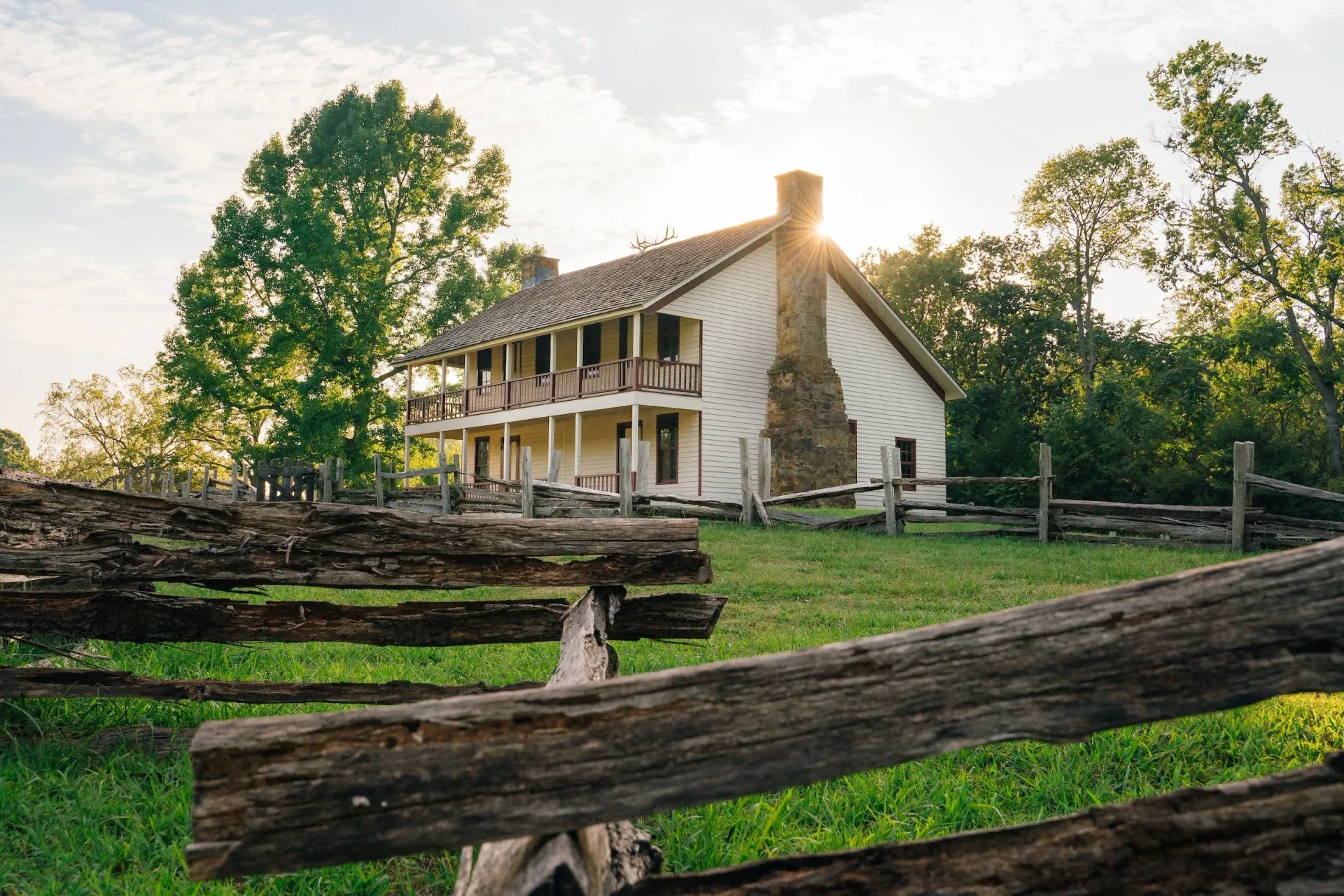 Rustic farmhouse with white siding behind a split-rail fence, surrounded by green trees at sunset.