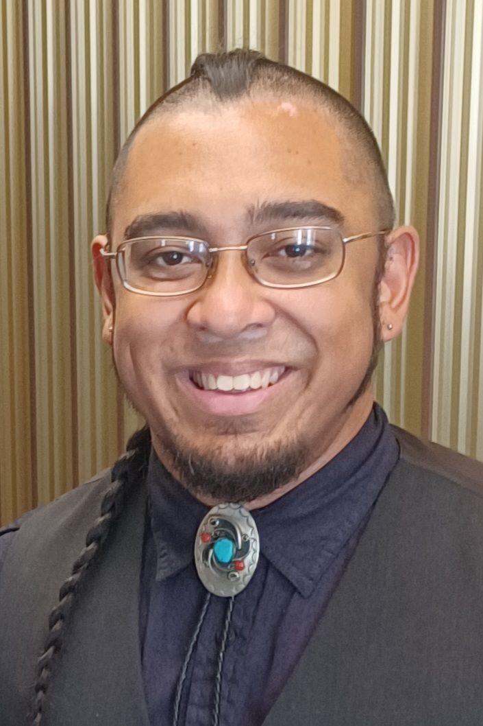 Smiling person in glasses and dark clothing with a decorative bolo tie, standing before striped curtains