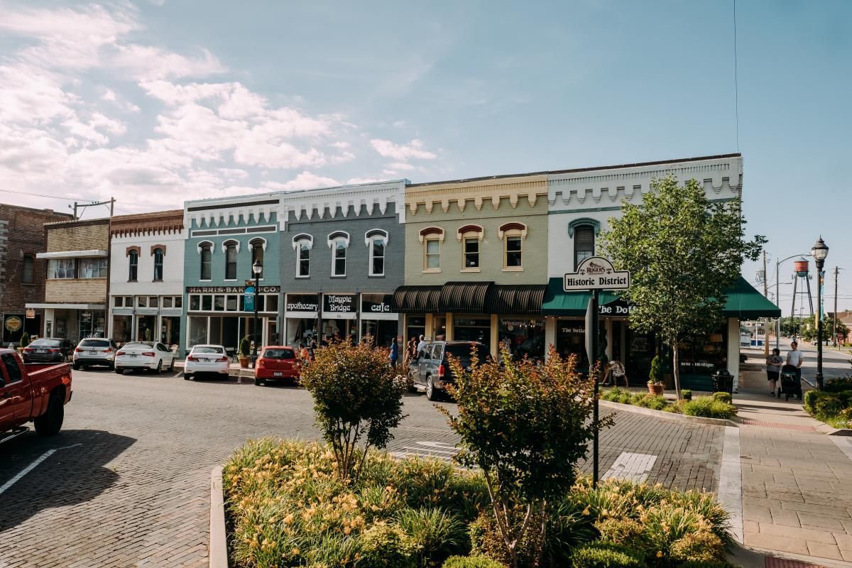 Historic downtown street with colorful storefronts, parked cars, and trees on a sunny day
