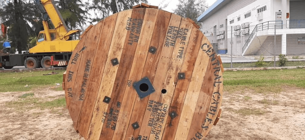 A large wooden cable spool on the ground with a crane and building in the background.