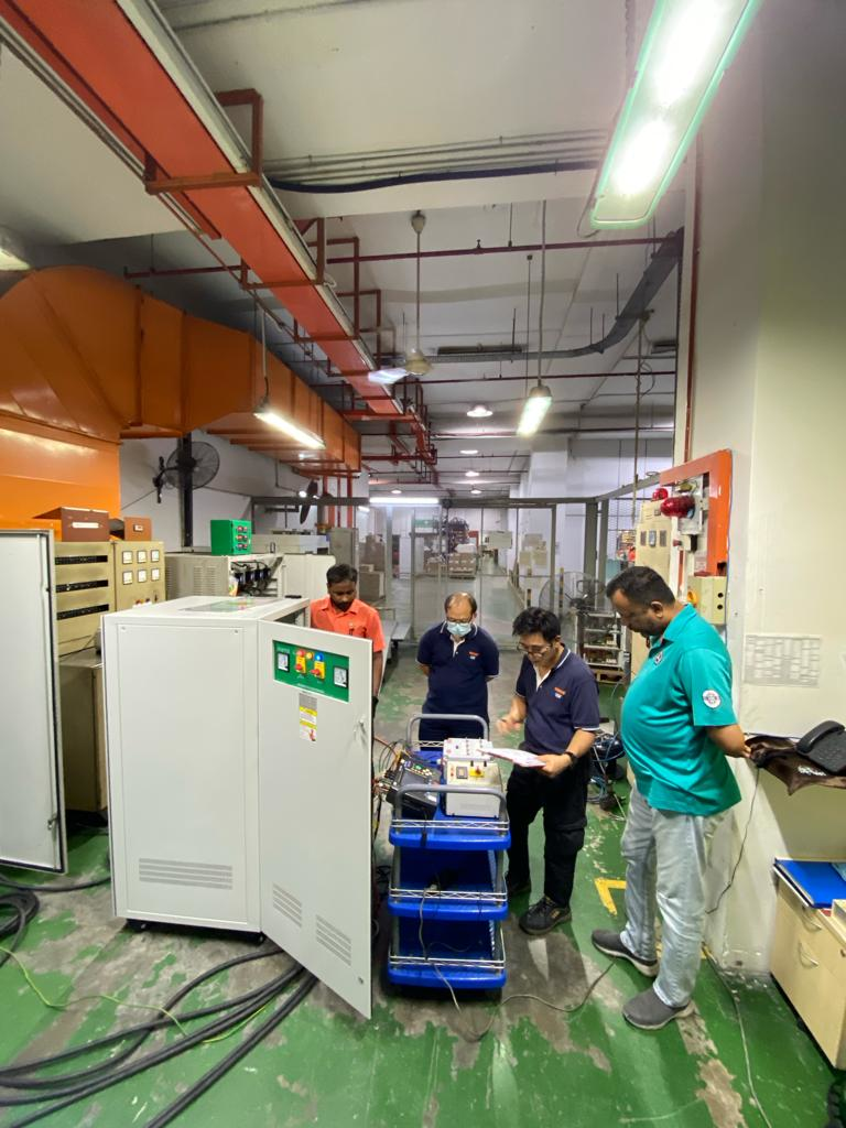 Four men in a factory setting inspecting equipment, a large white electrical unit in the foreground.