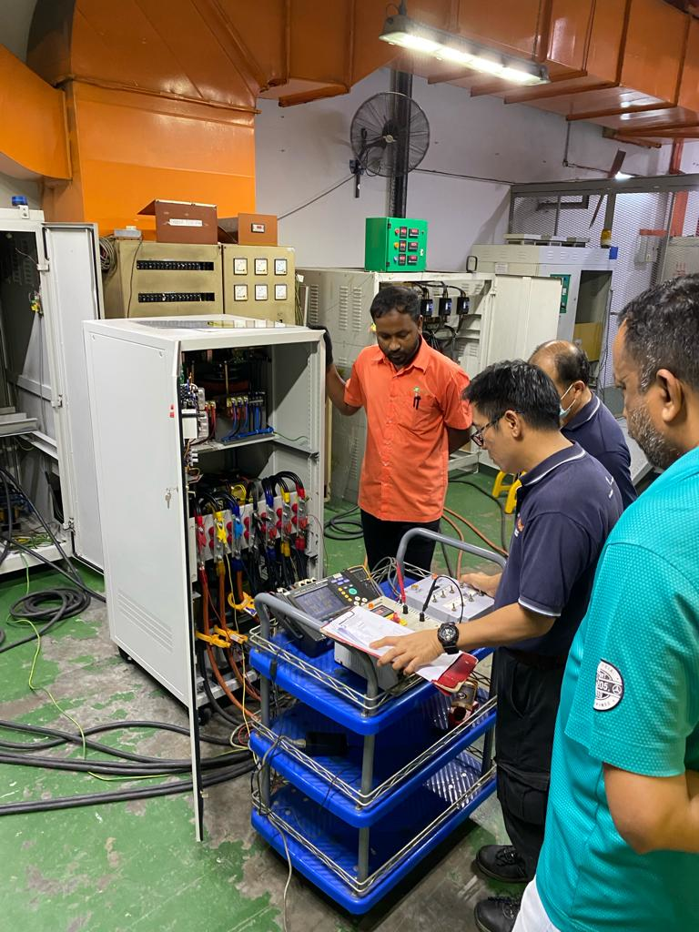 People examining electrical equipment in a factory setting. One person in orange shirt, others are looking on.
