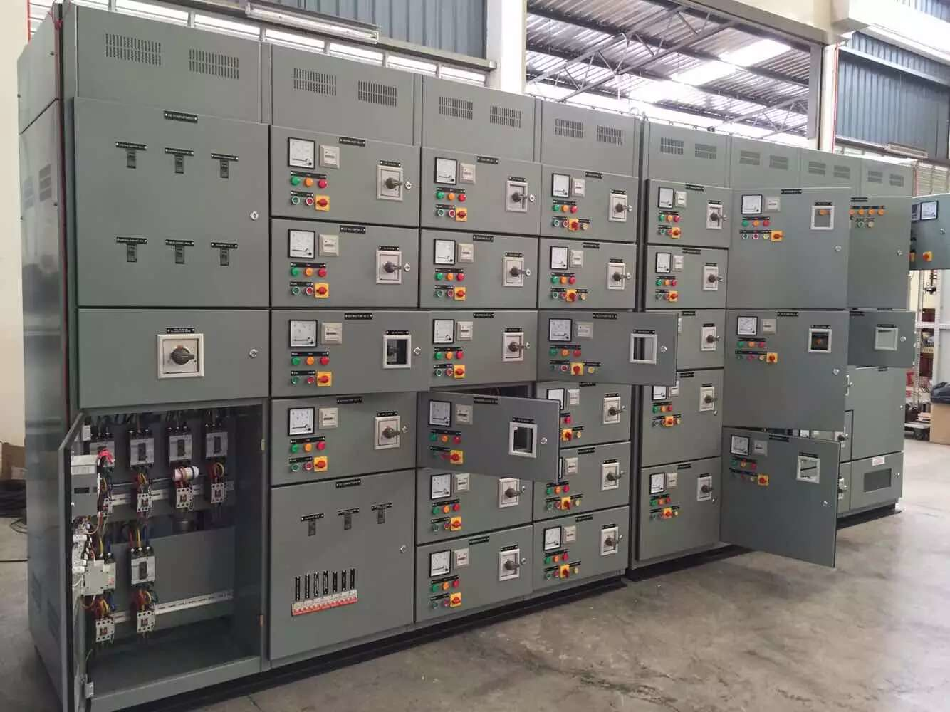 Gray electrical control panels with various switches, meters, and open doors in a factory setting.