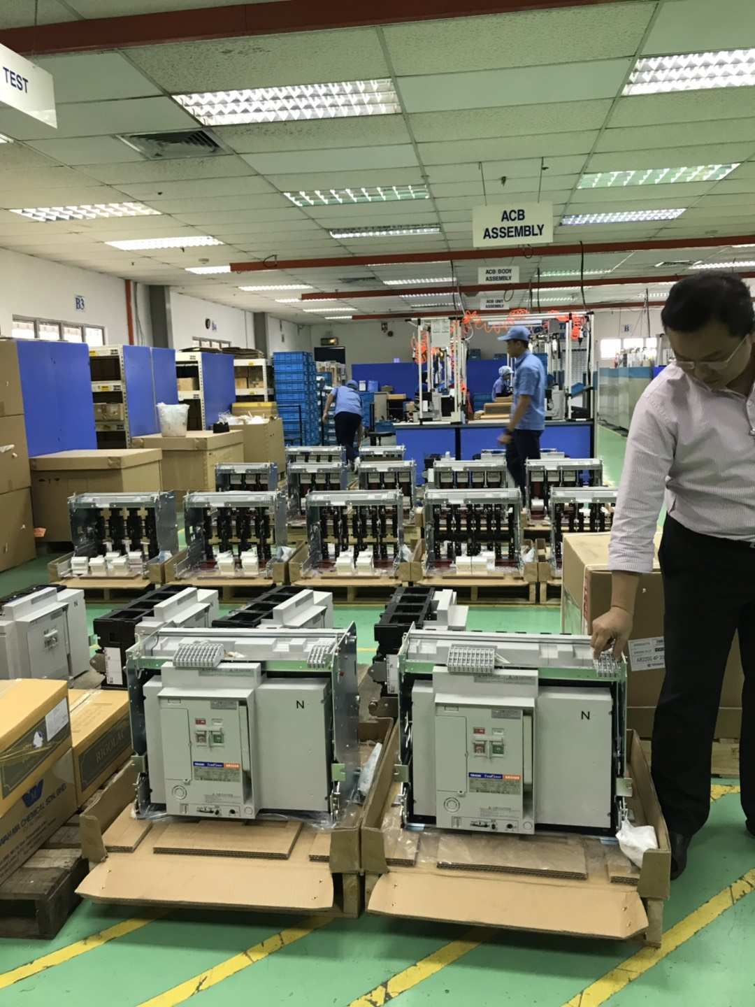 Man in factory examines electrical equipment. Multiple units are on pallets; workers in the background.