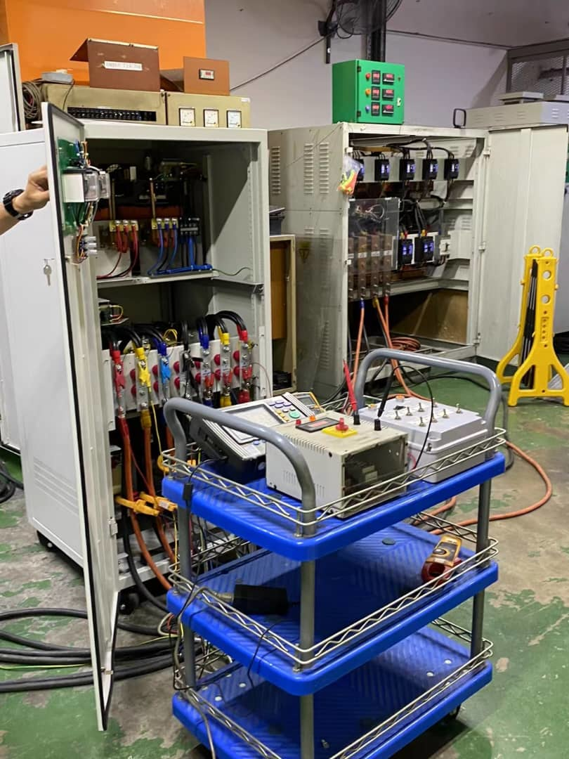 An open electrical cabinet with wires and components; a tool cart with equipment sits in front.