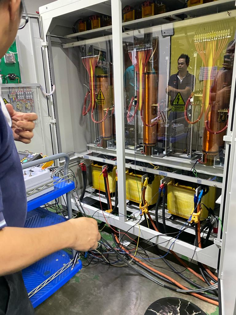 People examining electrical equipment with copper coils and transformers inside a cabinet; warning labels present.