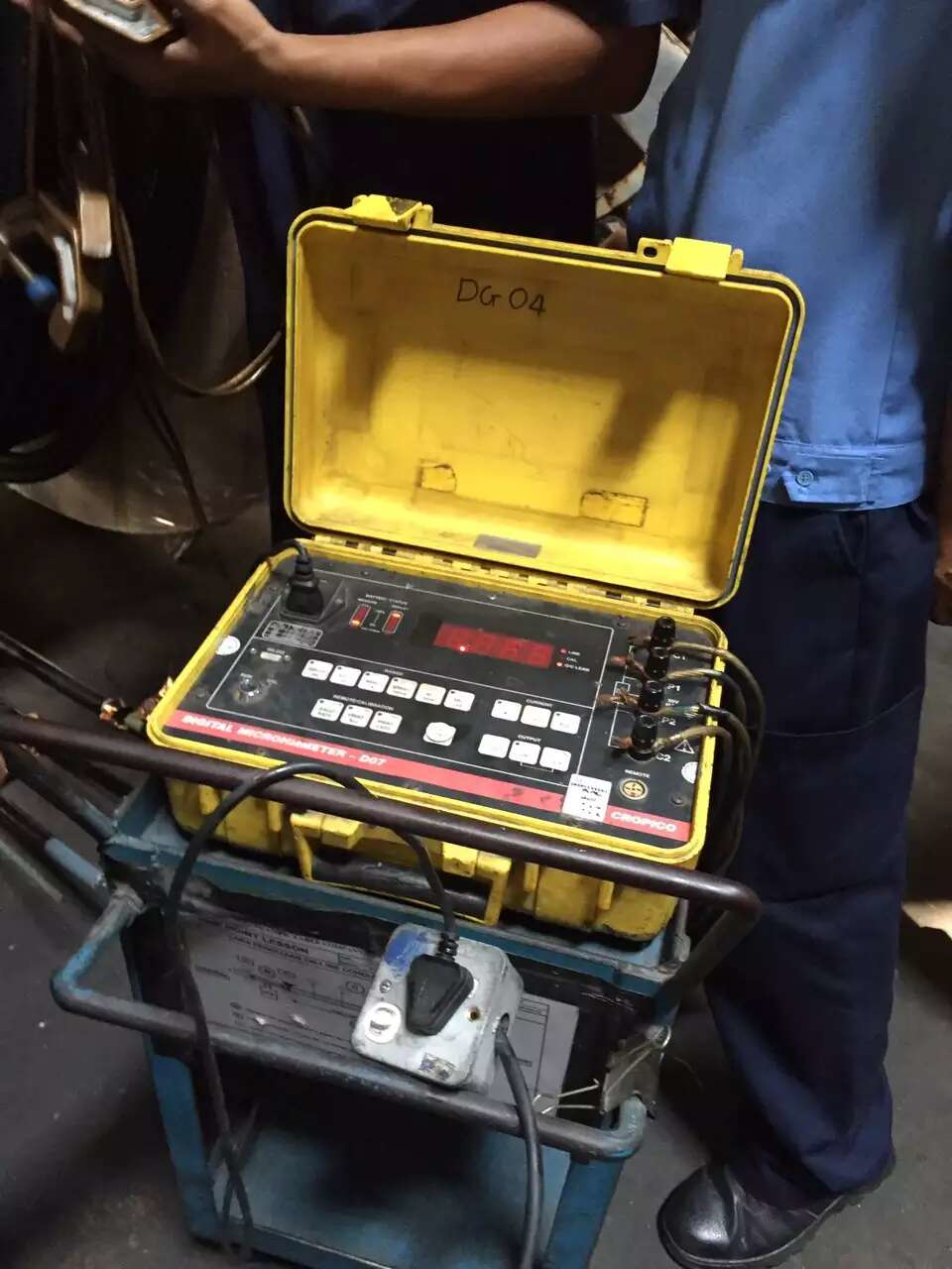 Yellow device with black cables on a cart, possibly for testing, used by workers in a shop.