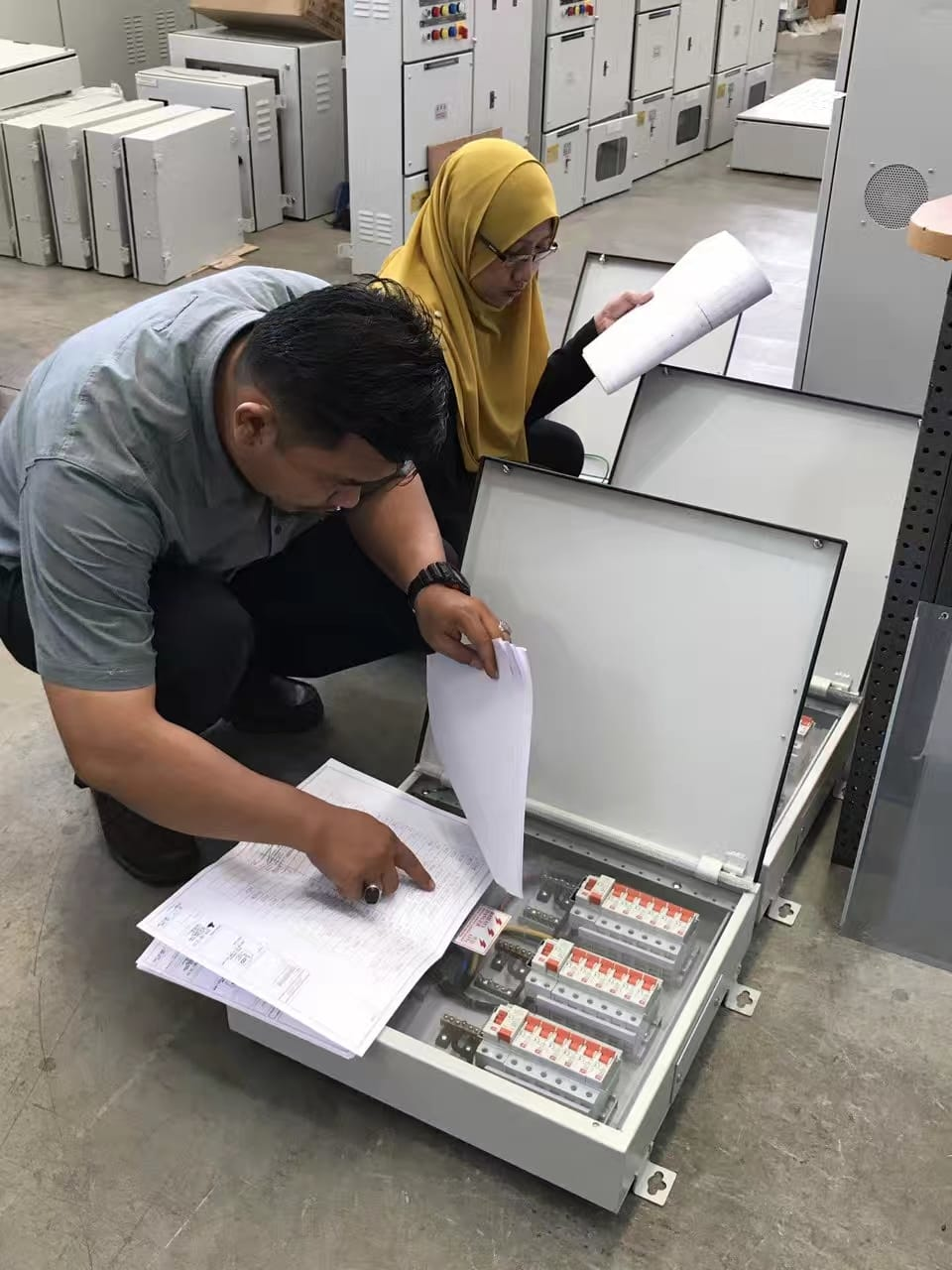 Two people inspecting electrical boxes with papers in a warehouse.