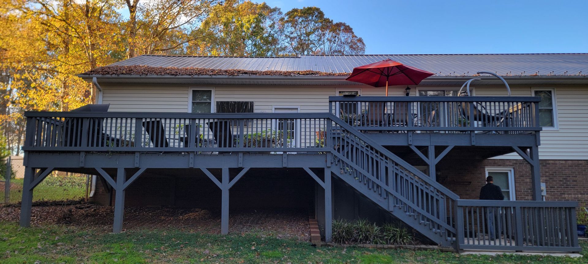 A two-tiered wooden deck painted dark gray on the back of a house. A red umbrella sits on the upper level. Trees with yellow leaves in the background.