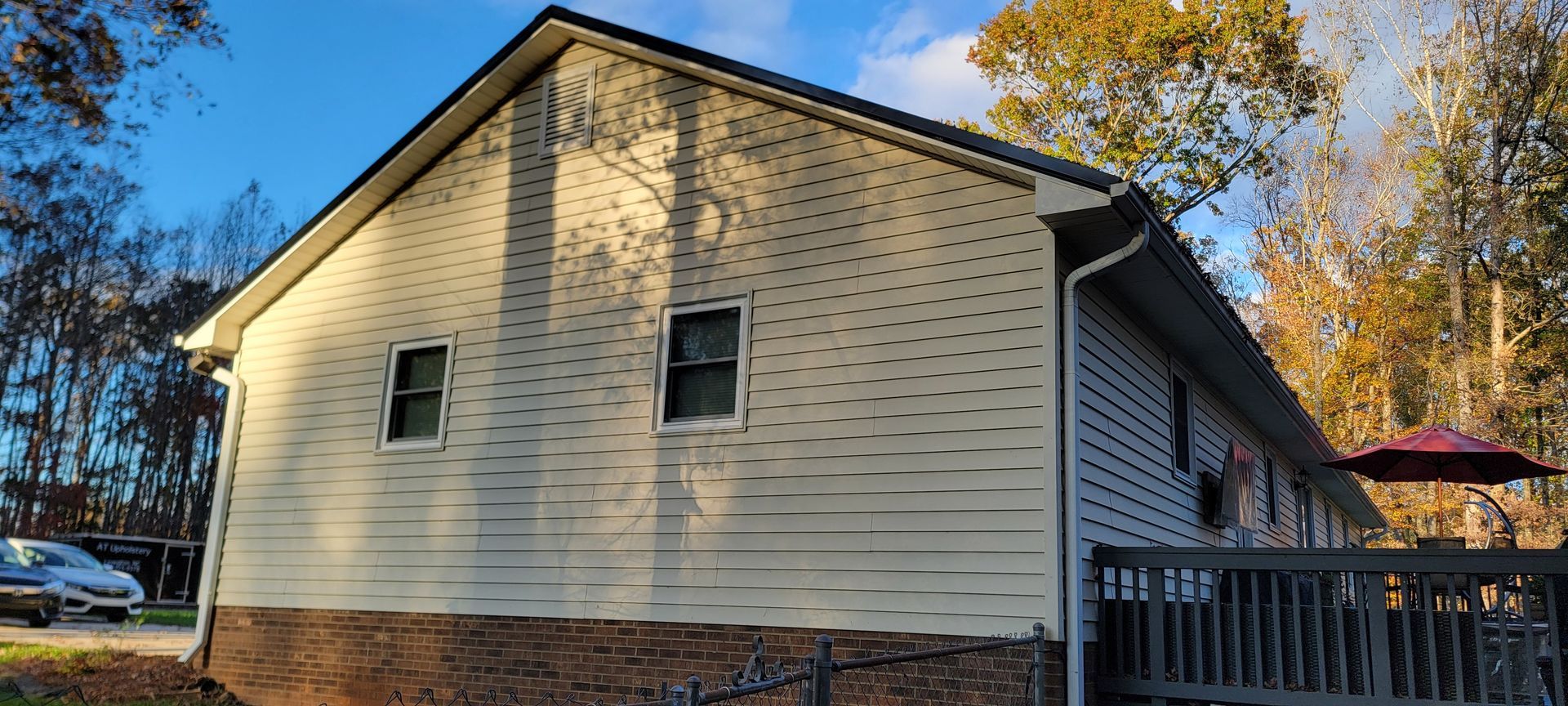 A two-story house with light-colored siding, two windows, and a dark roof. A deck with an umbrella is on the side.