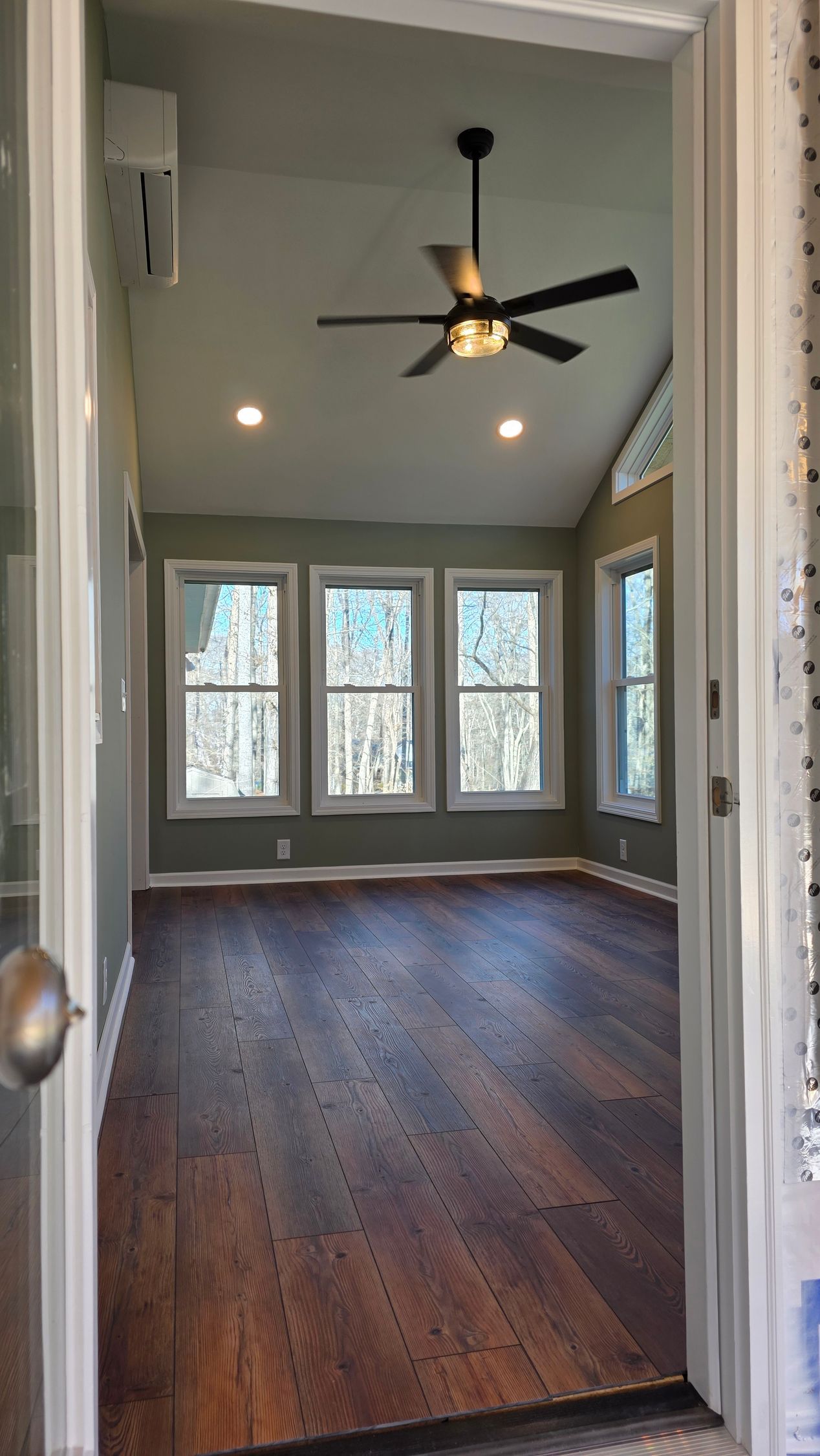 An interior shot of a room with hardwood floors, windows, and a ceiling fan, viewed from a doorway.