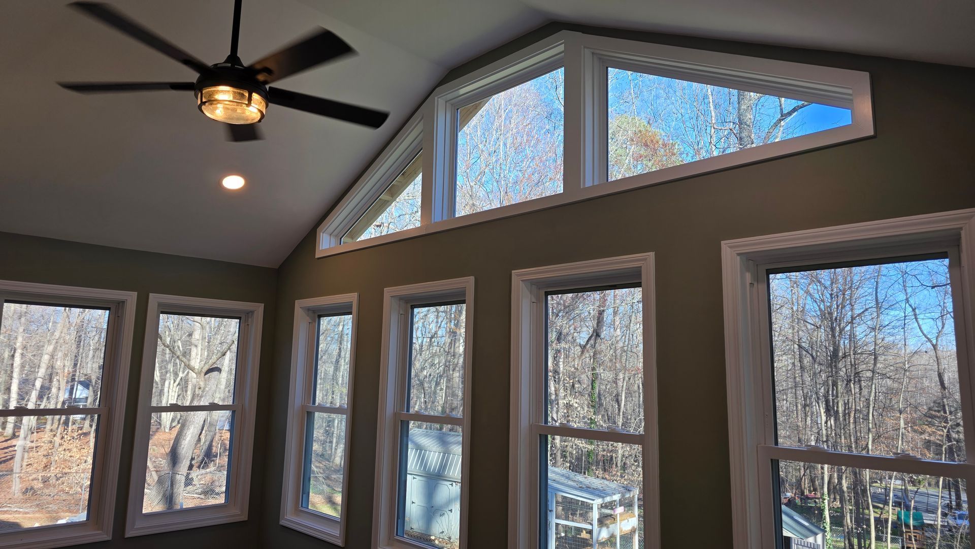 Interior view of a room with many windows, high angled ceiling, and a ceiling fan. Windows look out onto a forest.