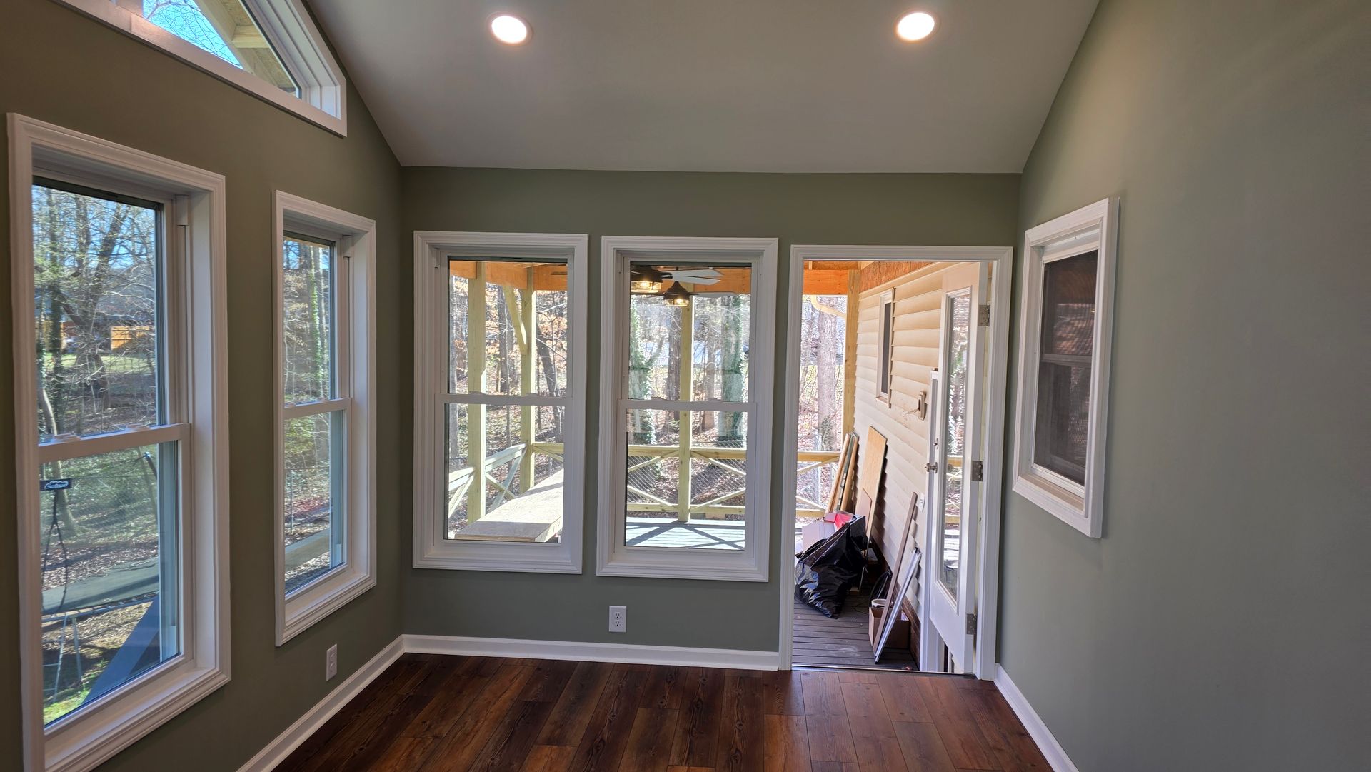 Sunroom with multiple windows and a door, featuring hardwood floors and light green walls. Trees are visible through the windows.