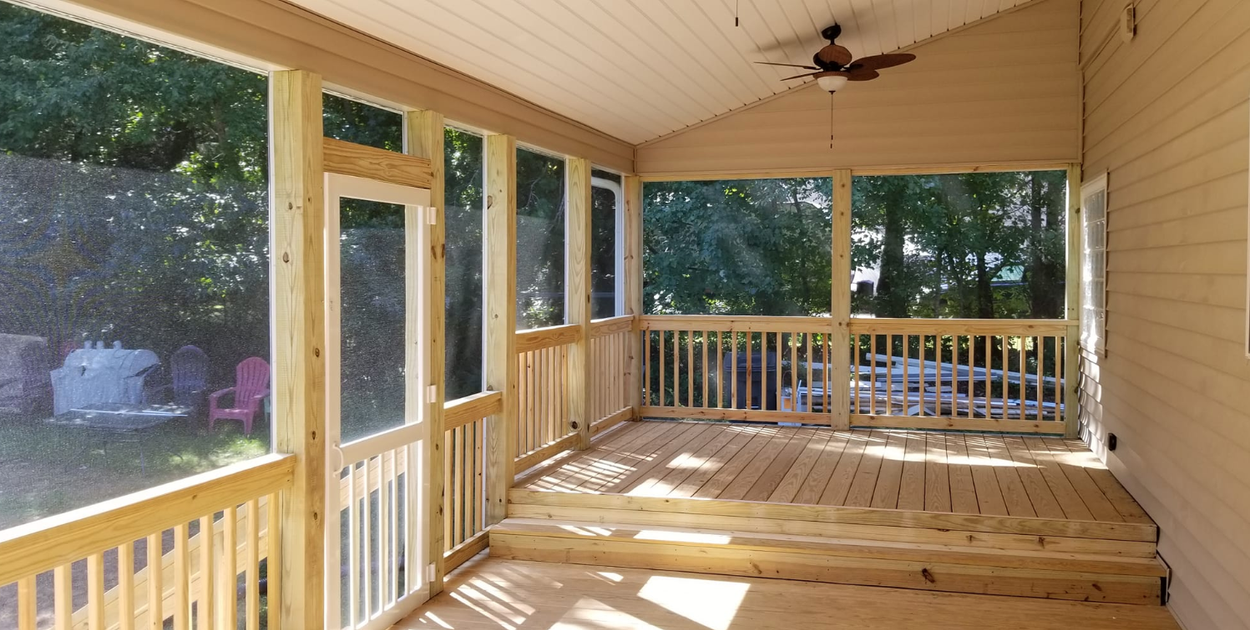 Screened porch with wooden deck, railings, and door. Trees and yard visible through the screens.