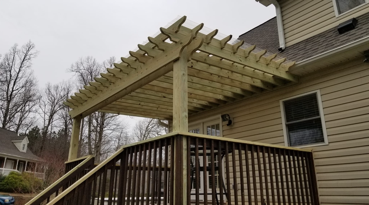 A wooden deck with a pergola attached to a beige house. Dark brown railings and steps lead up to the deck.