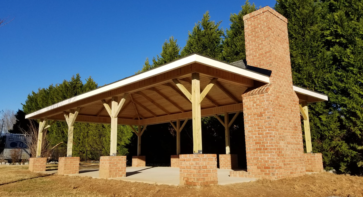 Outdoor pavilion with a brick chimney. The roof is white, supported by wooden beams and pillars on brick bases. A grassy area with trees in the background.